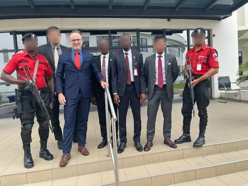 A group of five men in suits stand on steps with two armed security guards in red uniforms; all have blurred faces. They pose in front of a modern building with glass doors and a metal railing.