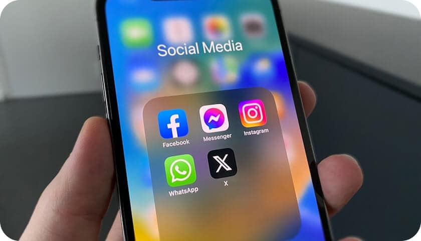 A hand holding a smartphone displaying a folder labeled "Social Media" with Facebook, Messenger, Instagram, WhatsApp, and X (formerly Twitter) app icons on the screen.