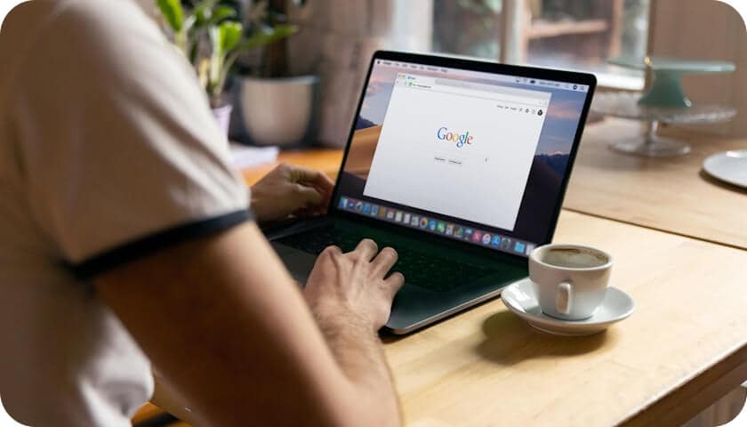 A person sits at a wooden table using a laptop with the Google homepage open; a cup of coffee and a plant are nearby, and sunlight streams in through a window in the background.