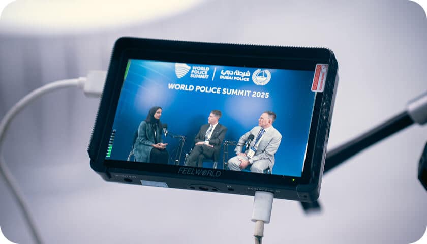 A close-up of a camera monitor displays three people seated on stage, engaged in discussion at the World Police Summit 2025, with event branding and logos visible on the screen.