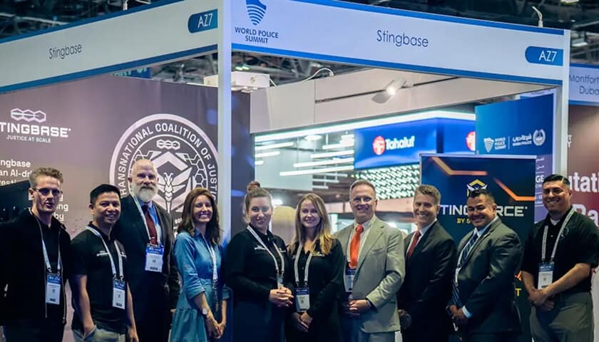A group of people in business attire pose and smile for a photo at a booth labeled "Stingbase" during the World Police Summit, with banners and displays visible in the background.