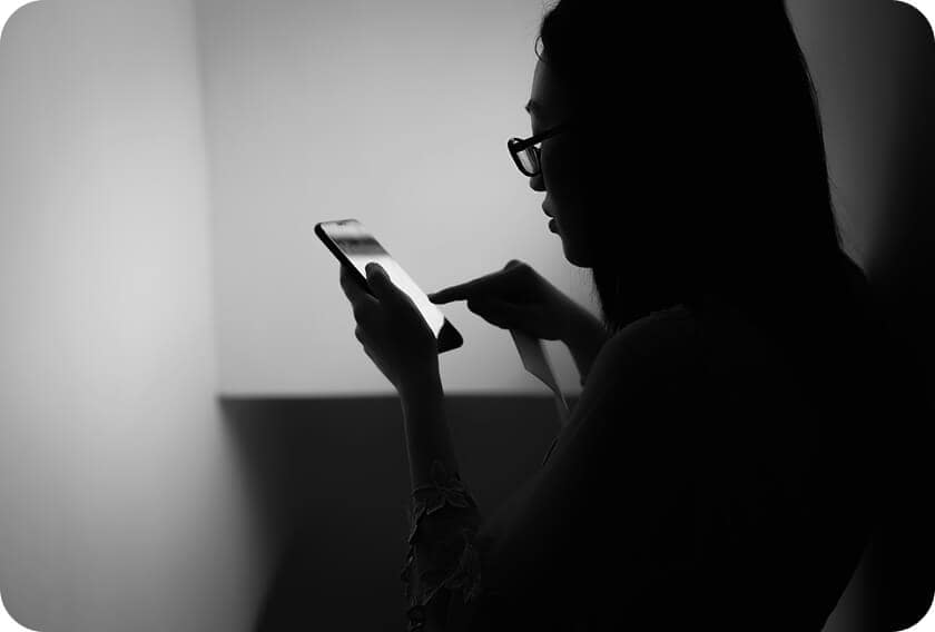 A person in silhouette wearing glasses uses a smartphone, touching the screen, with light illuminating their face and hand in an otherwise dark room.