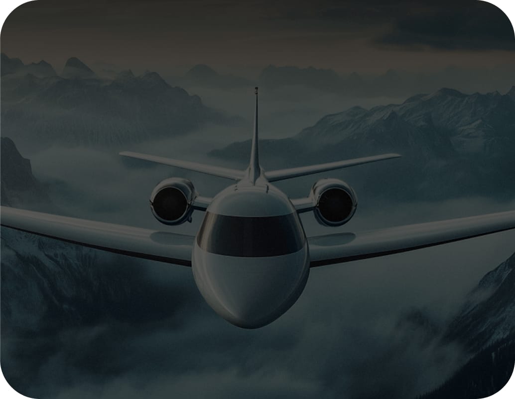 A jet airplane flies above misty mountains at dusk, with dramatic clouds and peaks in the background. The aircraft is shown head-on, dominating the center of the image.