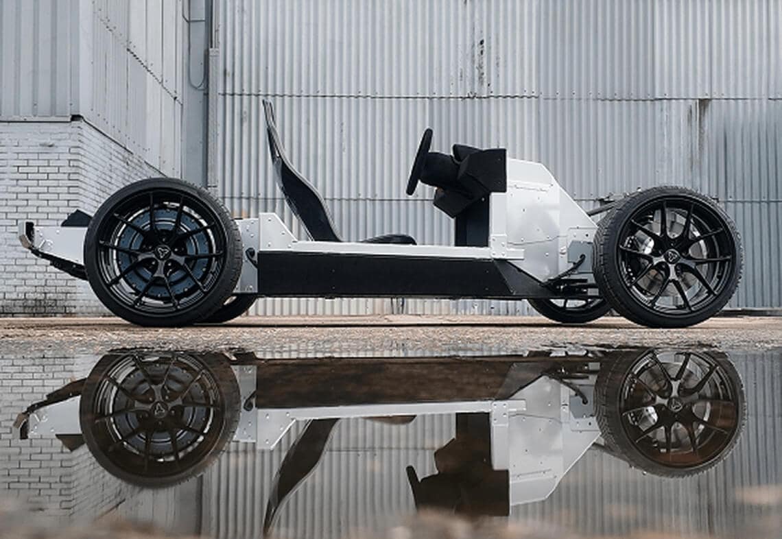 A minimalist electric vehicle chassis with exposed wheels, seat, and steering wheel is parked outdoors on concrete, reflected in a large puddle, with a corrugated metal building in the background.