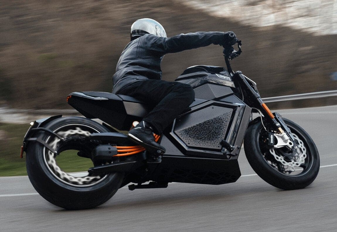 A person wearing a helmet and black leather jacket rides a black electric motorcycle at speed, leaning into a curve on a paved road, with blurred trees and hills in the background.