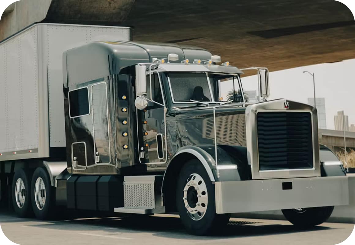 A shiny black semi-truck with chrome details and a large front grille is parked under an overpass in an urban area, with city buildings visible in the background.