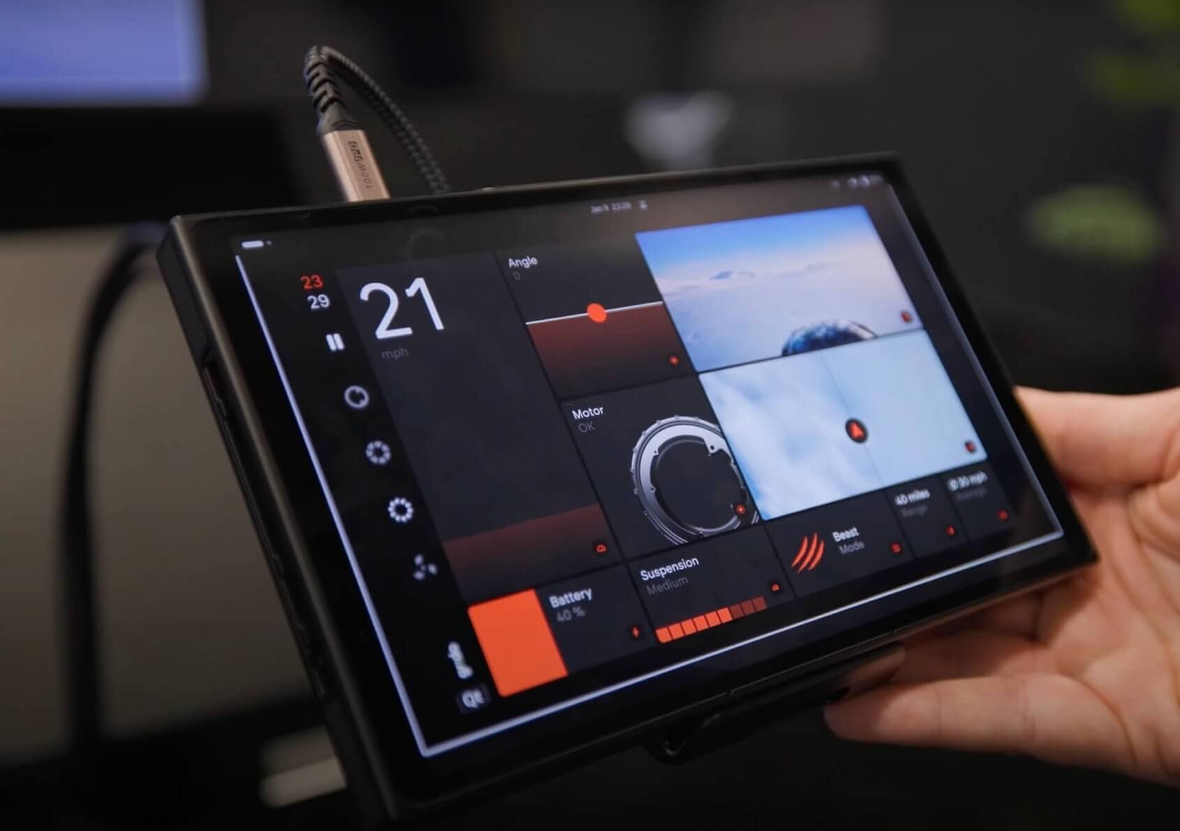 Close-up of a hand holding a black tablet device displaying a dashboard interface. The screen shows speed, angle, battery life, and other technical metrics with various dials and indicators against a dark background.