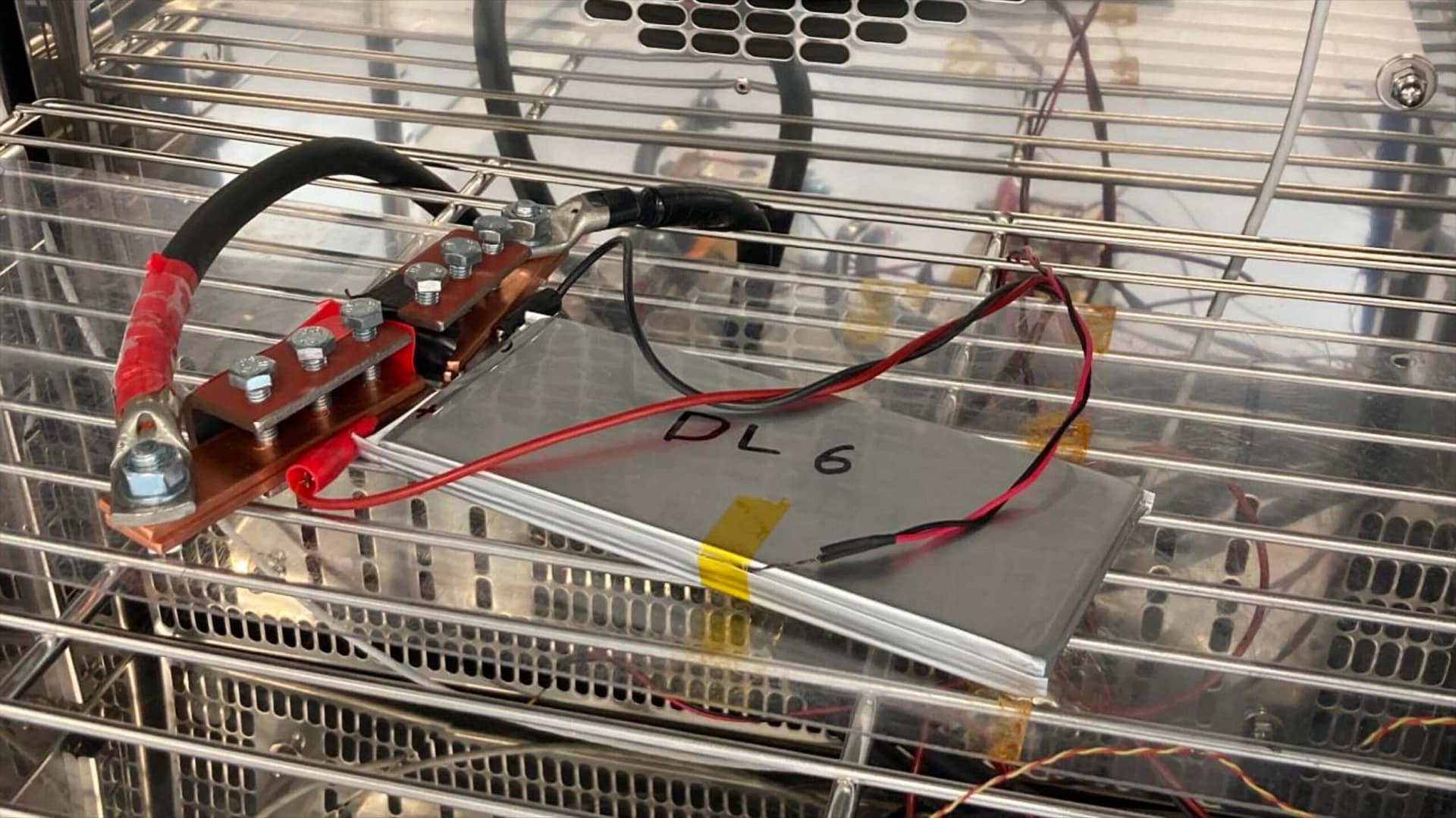 A silver battery cell labeled “DL 6” sits on a metal rack with red and black wires attached, inside a metal enclosure with vented walls and bolts.