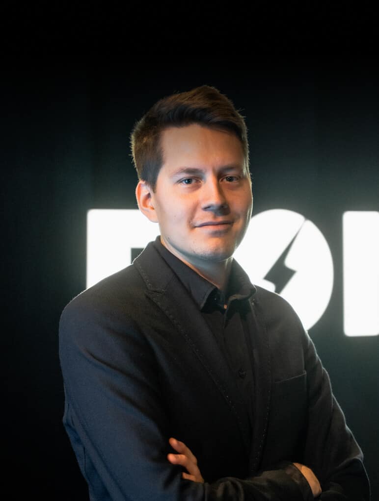 A person in a dark suit stands confidently with arms crossed against a dark background with a partially visible illuminated logo. The lighting highlights the person's face and the formal attire creates a professional appearance.
