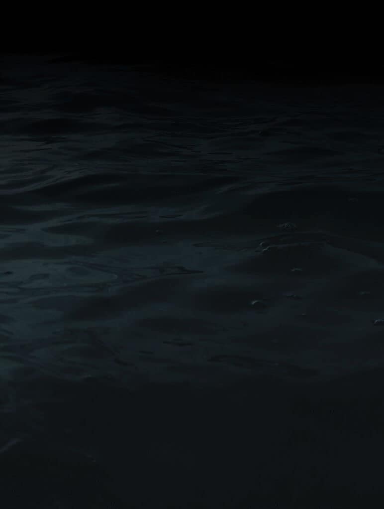 A dark, mysterious body of water with gentle ripples and subtle reflections under a dim light, creating a tranquil yet enigmatic atmosphere. The surface appears calm, and the background fades into black.