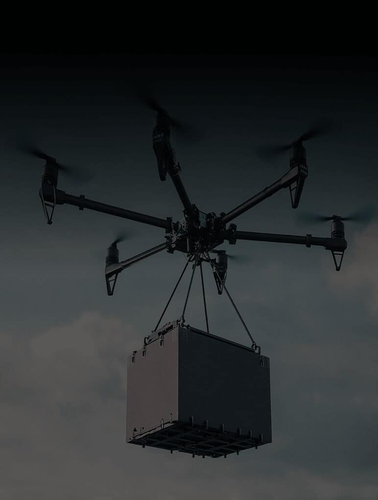 A drone with six rotors is carrying a large, rectangular package suspended below it against a cloudy sky.