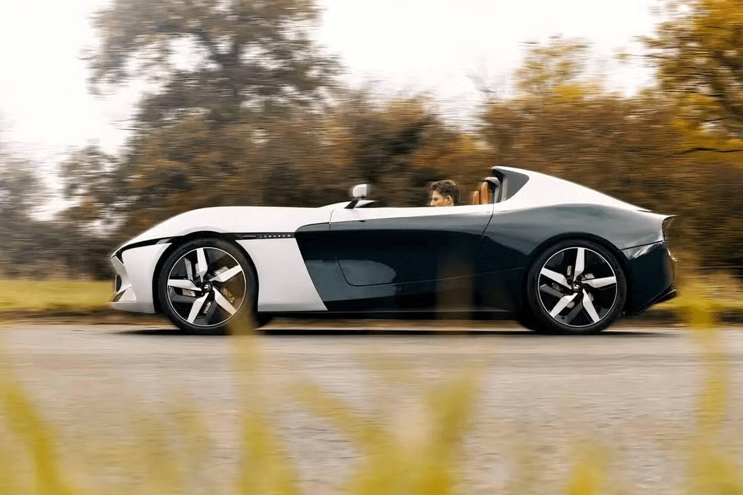 A person drives a sleek, modern, two-tone convertible sports car on a road with blurred autumn trees in the background and green grass in the foreground.