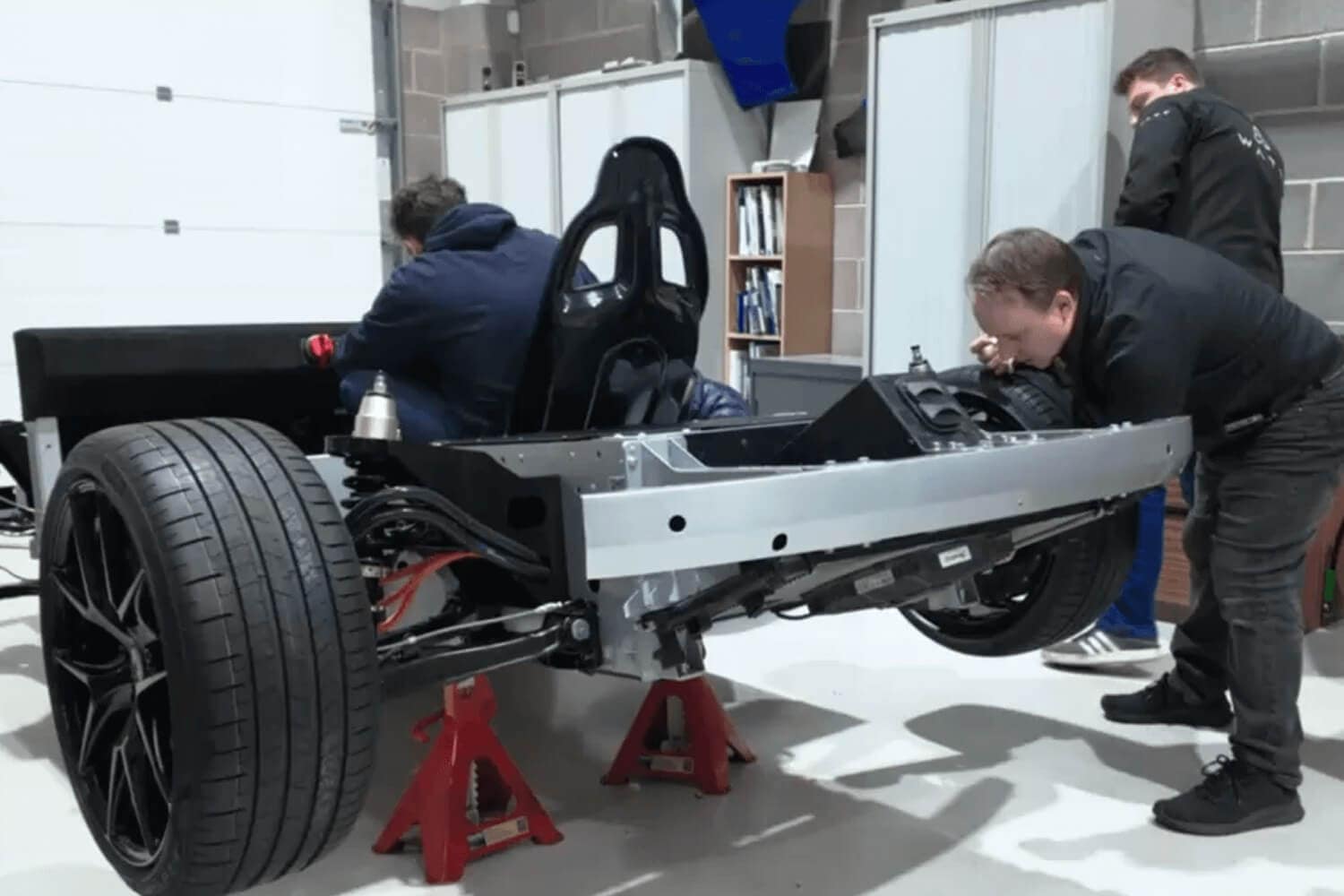 Two people work on a car chassis elevated on red jack stands in a garage or workshop. The car has large tires and a visible black racing seat, with tools and shelves in the background.