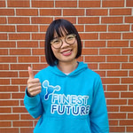 Bambi Dang A person with short black hair and glasses smiles and gives a thumbs up. They are wearing a bright blue hoodie that says "FINEST FUTURE," standing in front of a brick wall.