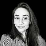 Emisa Lehtinen Black and white portrait of a woman with long hair, smiling softly. She wears a striped shirt and looks directly at the camera, against a solid black background.