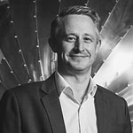 Dan Walmsley Esox portrait A man in a suit smiles at the camera, standing in front of a metallic, riveted, fan-shaped background. The photo is in black and white.