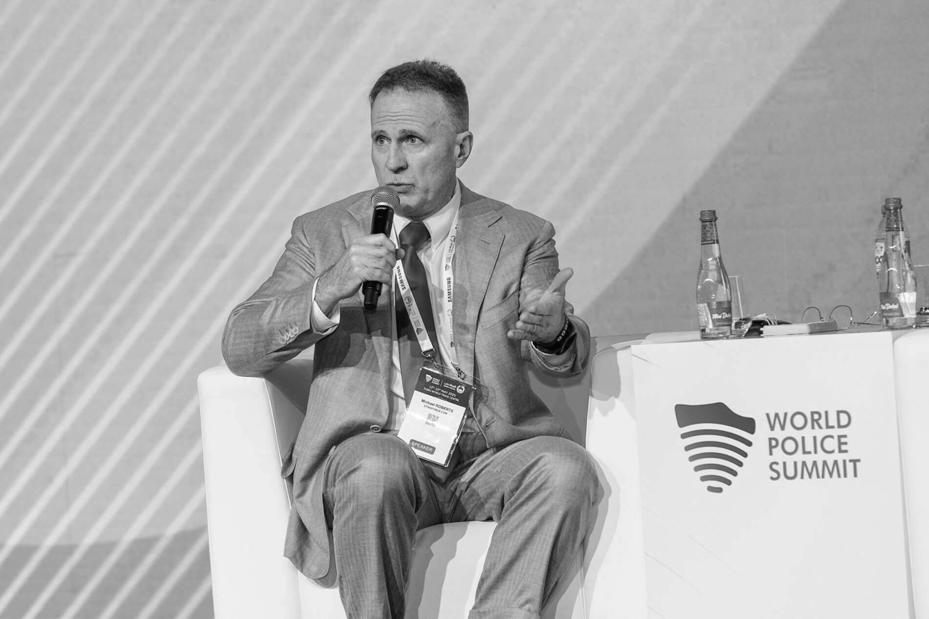 Michael Roberts A man in a suit speaks into a microphone while seated on a stage at the World Police Summit. Two water bottles and event signage are on a table beside him.