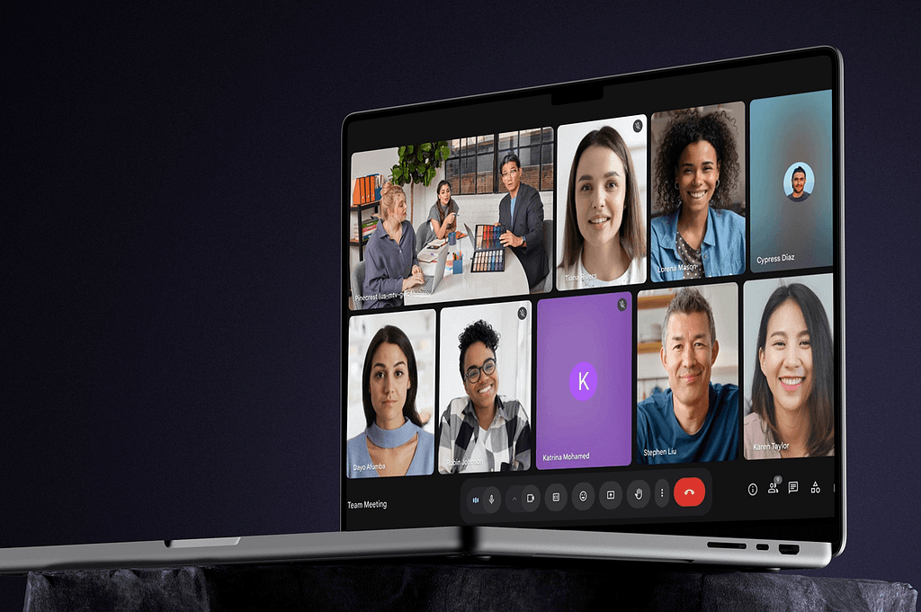 A laptop displays a virtual meeting with nine diverse participants, some with cameras on and others using profile pictures, set against a dark background. Meeting controls are visible at the bottom of the screen.
