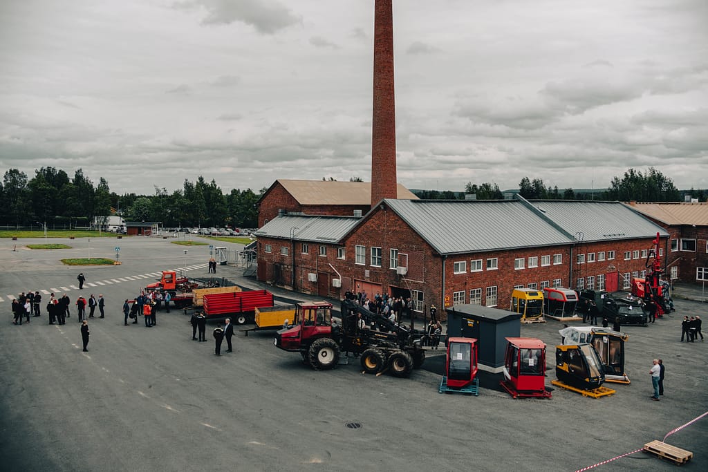 A group of people gather around various construction vehicles and machinery parked in front of a large red-brick industrial building with a tall chimney on a cloudy day.