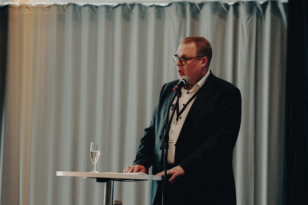 A man in a dark suit and glasses speaks into a microphone at a podium with a glass of water on it, in front of light-colored curtains.
