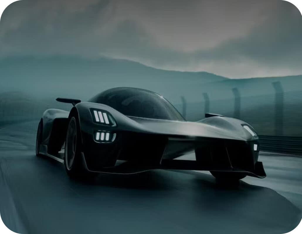 A sleek, black sports car drives on a wet racetrack at dusk, with mountains and a cloudy sky in the background.