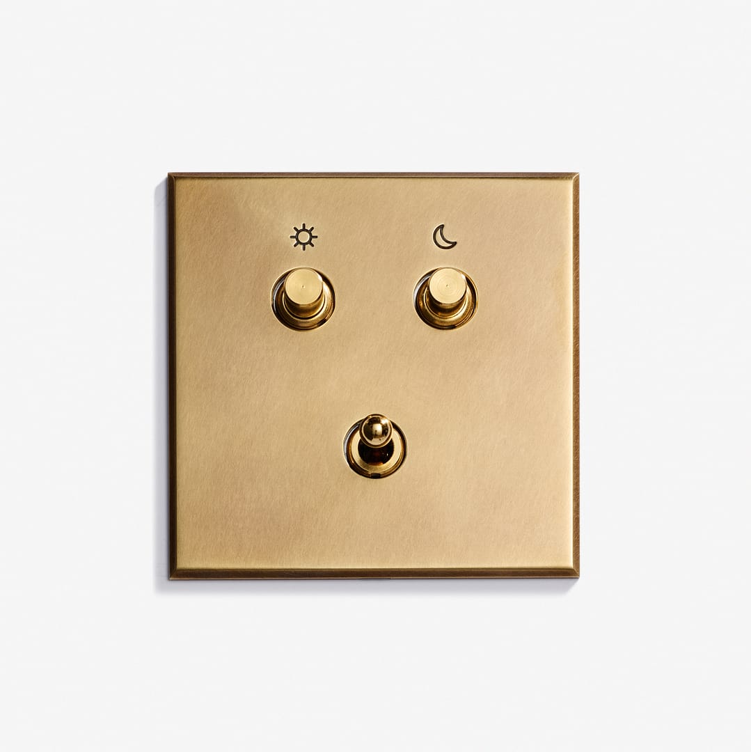 A gold-colored Classique Keypad 82 x 82 light switch panel with three toggle switches: the top left marked with a sun symbol, the top right with a moon, and one unmarked switch centered below them.