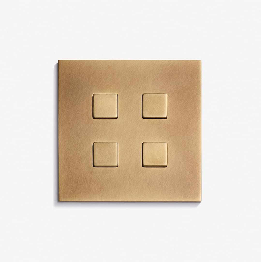 A Damier Keypad 82 x 82 brass light switch plate features four evenly spaced square buttons, mounted on a plain white wall.
