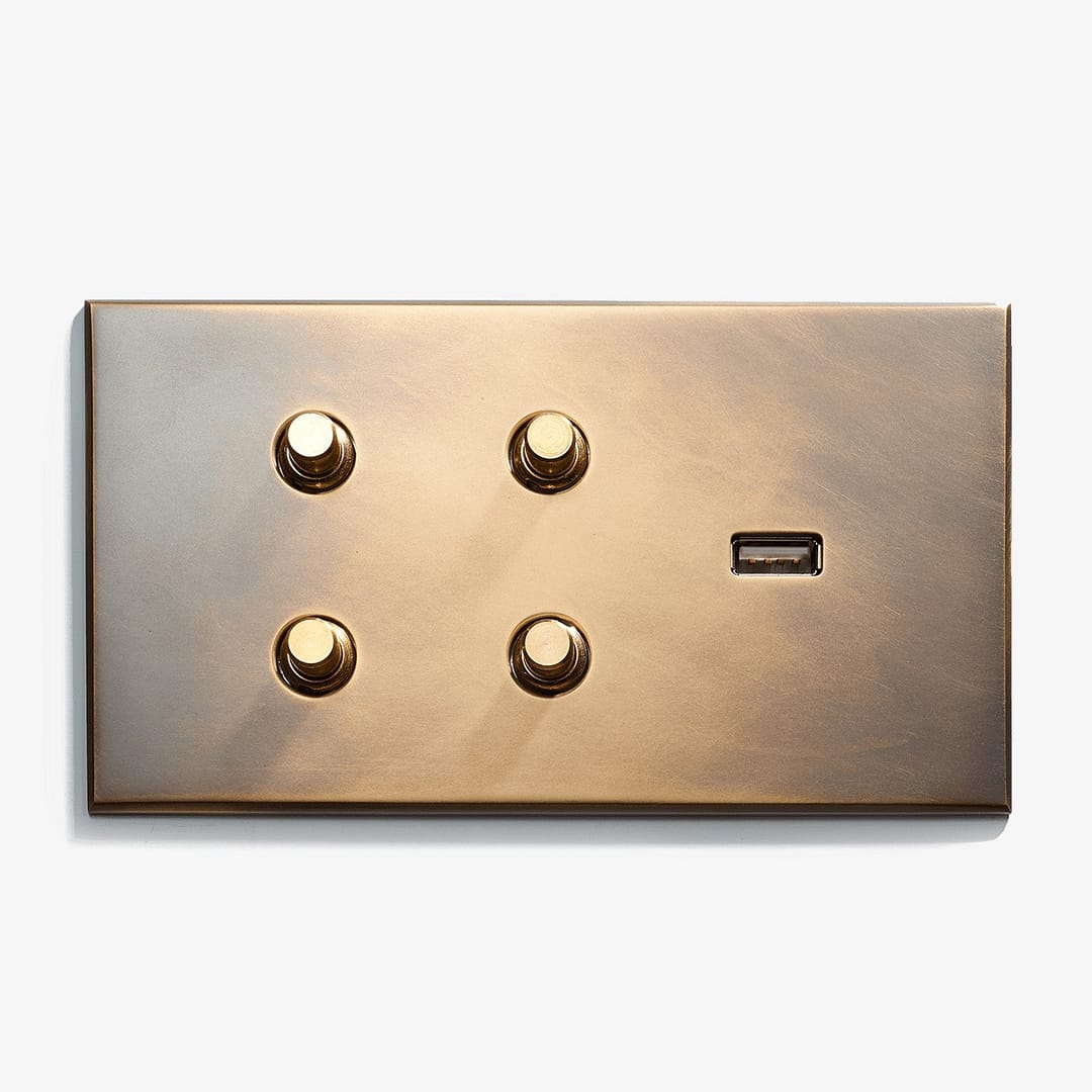 A burnished gold wall plate featuring four round buttons and a usb port, illuminated by a bright light casting soft shadows, isolated on a white background.