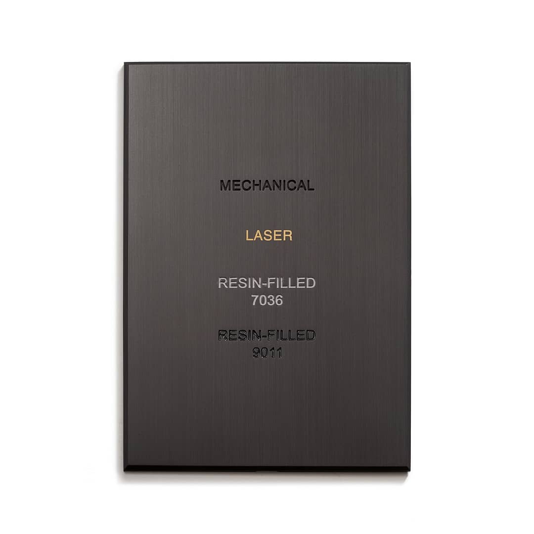 A close-up of a dark metallic plaque displaying different types of text inscriptions. The plaque has the words "MECHANICAL," "LASER," "RESIN-FILLED 7036," and "RESIN-FILLED 9011" engraved, showing different engraving techniques.