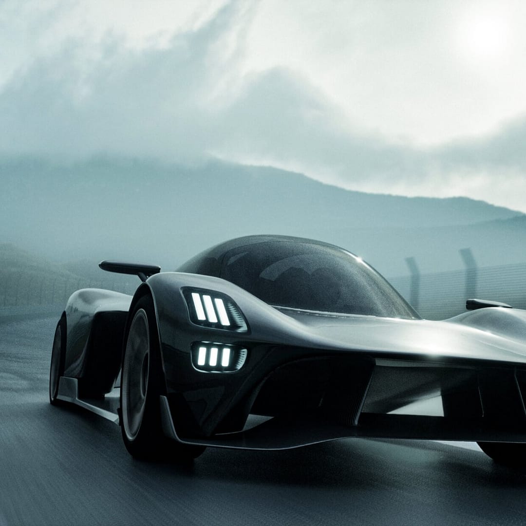 automotive-sbs A sleek, futuristic sports car speeds along a racetrack. The car features aerodynamic curves, prominent air intakes, and glowing headlights. A misty mountain range is visible in the background under a cloudy sky.