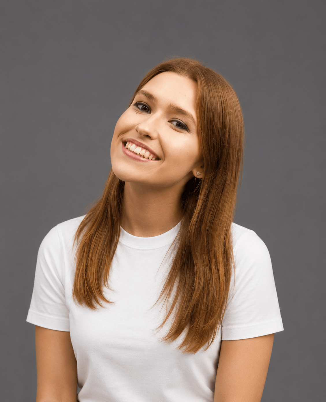 A young woman with straight, reddish-brown hair smiles, tilting her head slightly. She wears a plain white t-shirt and is posed against a neutral gray background.