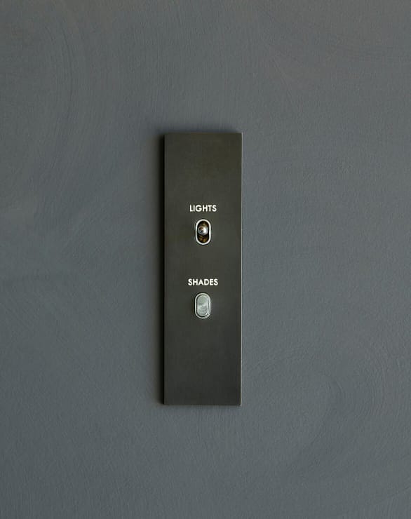 A minimalist wall panel with two buttons labeled "LIGHTS" and "SHADES" is mounted on a plain gray wall.