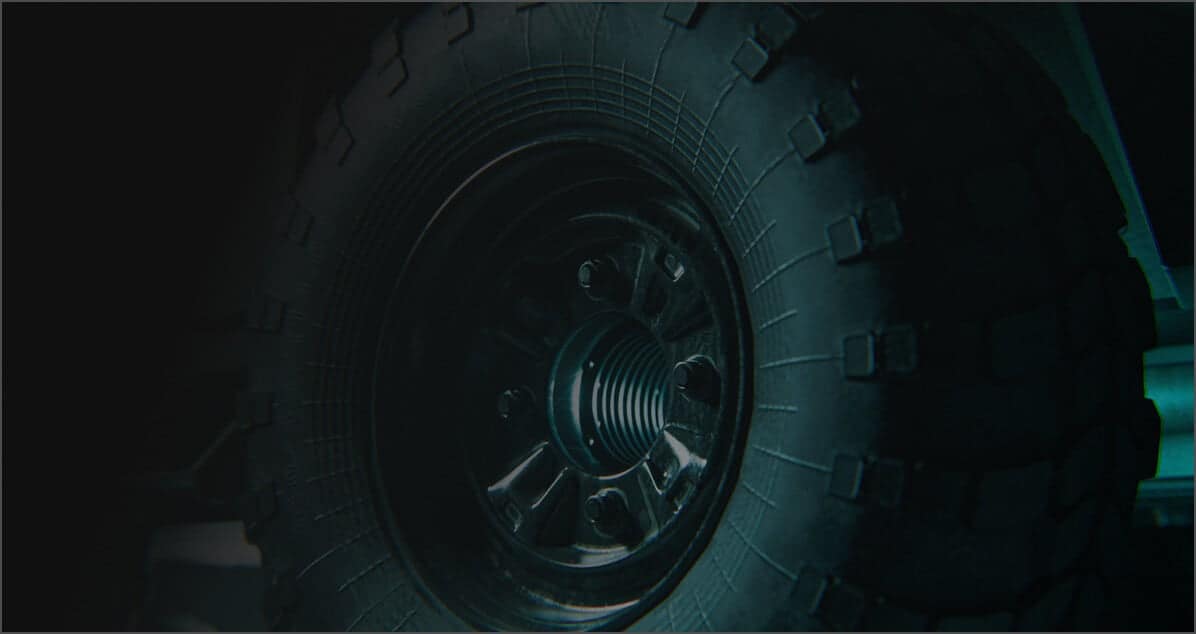 Close-up view of a rugged, off-road vehicle tire with a dark rim, showing detailed tread patterns and bolts. The image is dimly lit, emphasizing the tire’s texture and sturdy construction.
