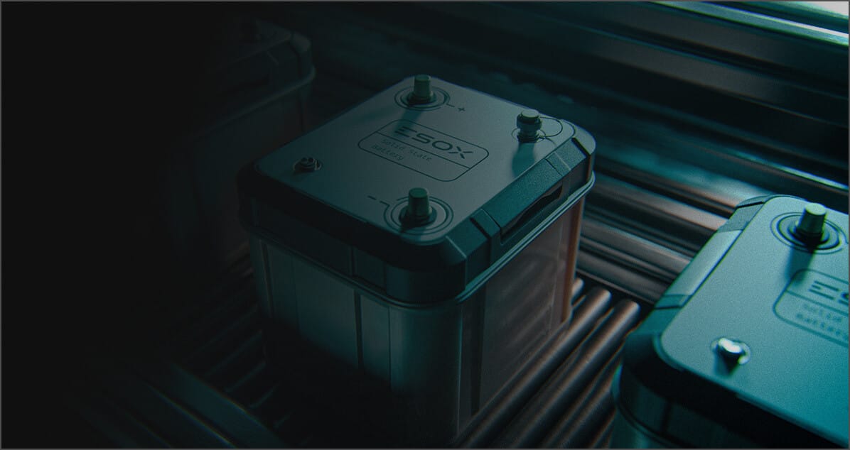 A close-up of a rectangular battery with labeled terminals, sitting on a metal conveyor belt. The lighting is dim, highlighting the battery's details and texture, with additional batteries partially visible in the background.