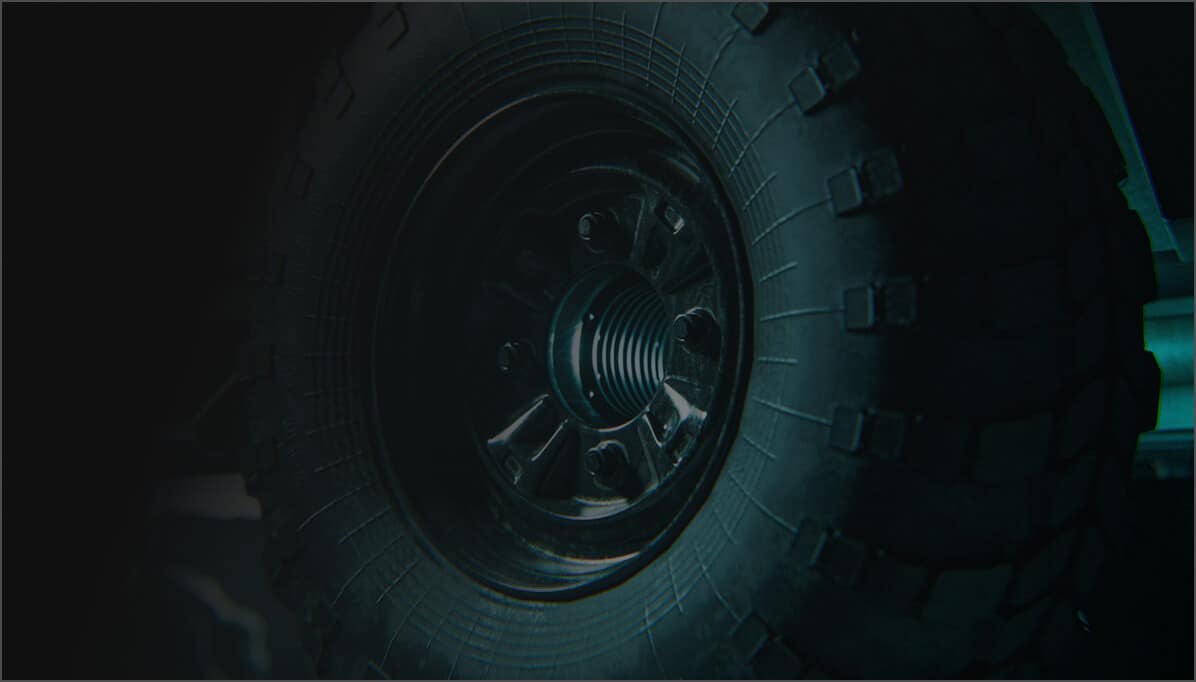 Close-up of a rugged, black off-road tire with deep treads and a metal wheel rim, partially illuminated with a greenish-blue light against a dark background.