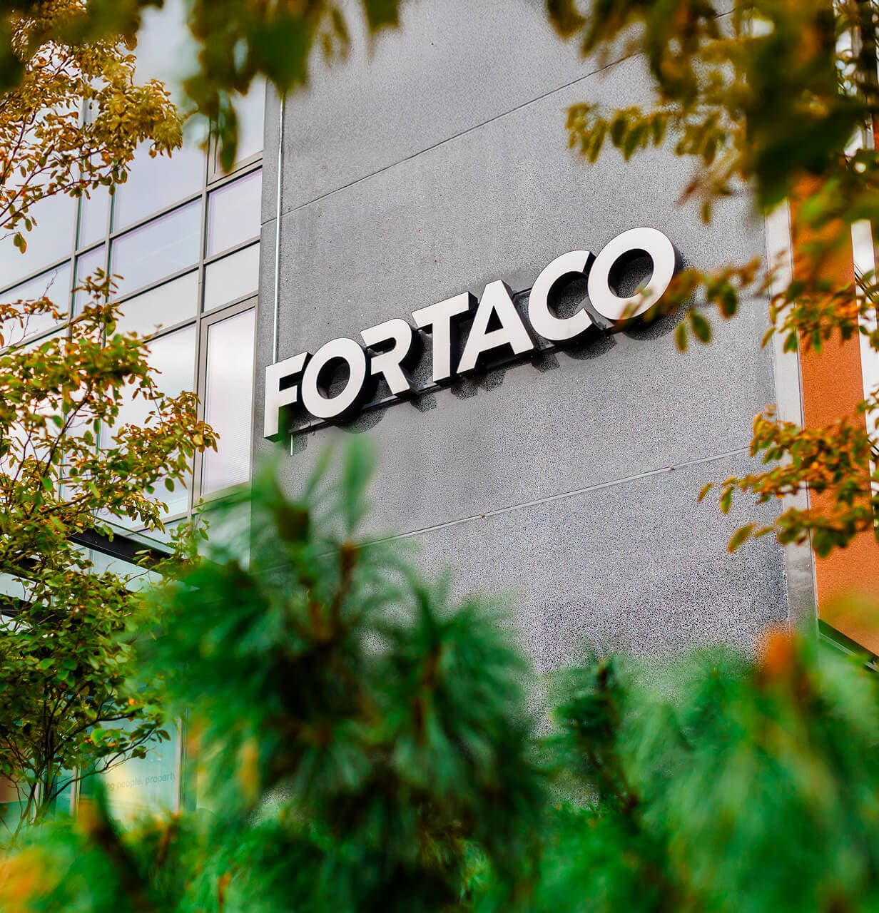 The exterior of a modern building with large windows and the word "FORTACO" displayed in bold white letters on a dark wall, partially framed by green trees and foliage in the foreground.