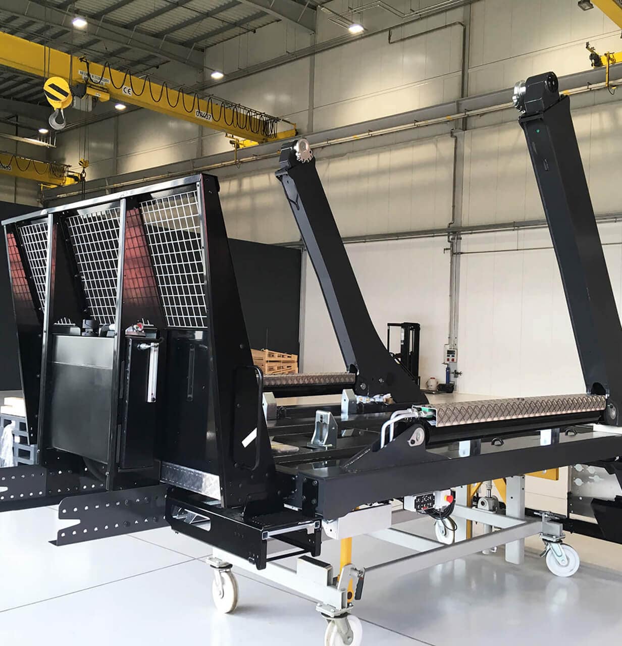 A large industrial conveyor system with black metal frames and rollers is set inside a spacious, well-lit factory with yellow overhead cranes and white walls and floors.