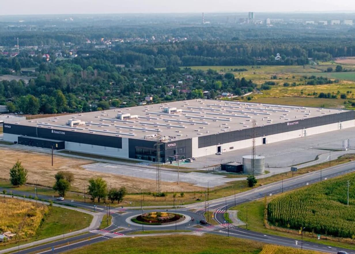 A large, modern industrial warehouse with a flat roof sits amid open fields and near a roundabout, with a distant view of trees, buildings, and a hazy skyline in the background.