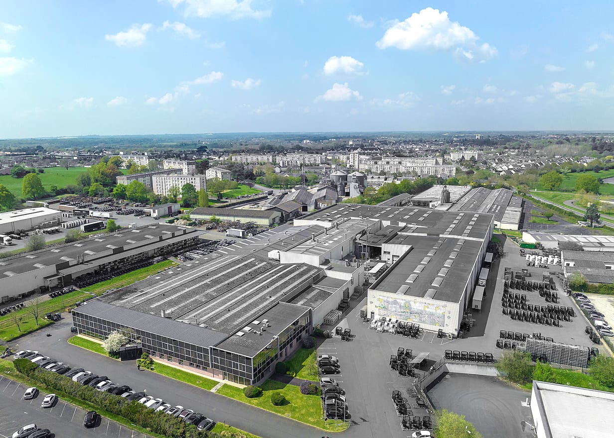 Aerial view of a large industrial complex with multiple long buildings, parking areas, and surrounding greenery; a cityscape stretches out in the background under a partly cloudy sky.