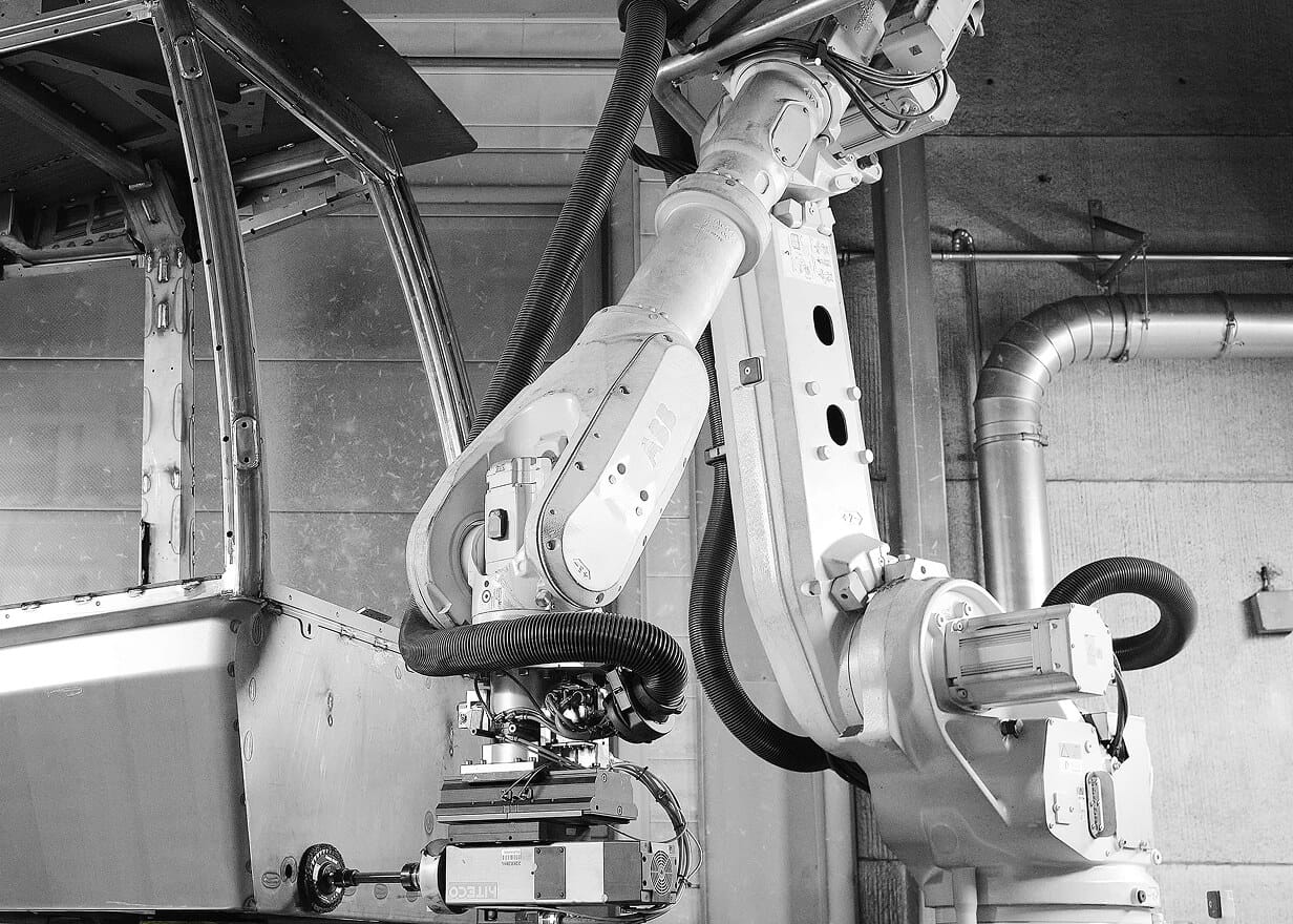 A close-up, black-and-white image of an industrial robotic arm with mechanical joints and cables, operating near a metal structure in a factory setting.