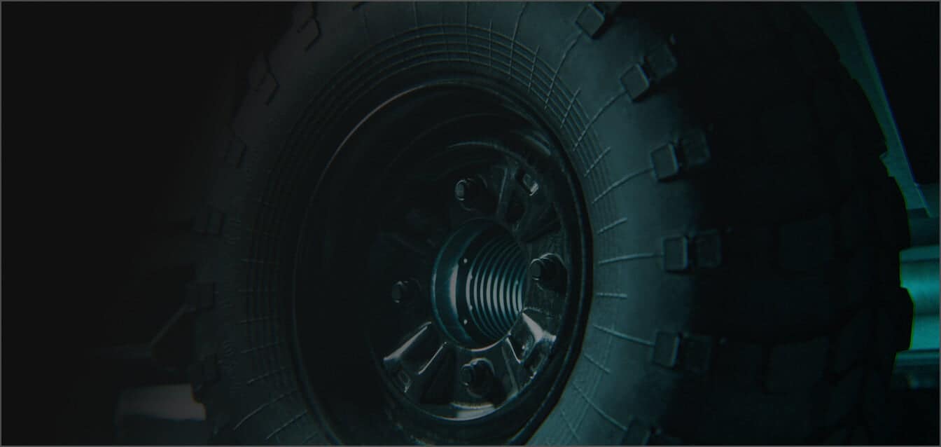 Close-up view of a rugged, black off-road vehicle tire with deep treads and a metallic wheel rim, shown in dim lighting.