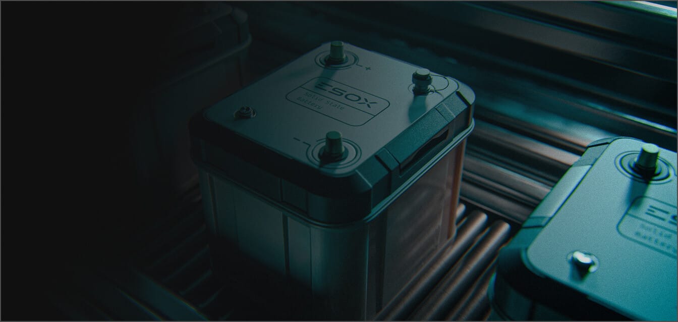 A close-up of a rectangular battery with labeled terminals, placed on a metal surface next to other similar batteries in a dimly lit environment.