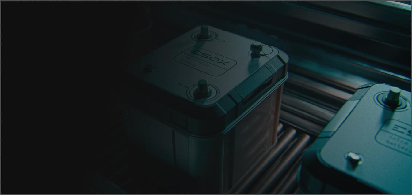 Close-up of two dark-colored batteries with visible terminals and labels, placed side by side on a metal surface with dim lighting.