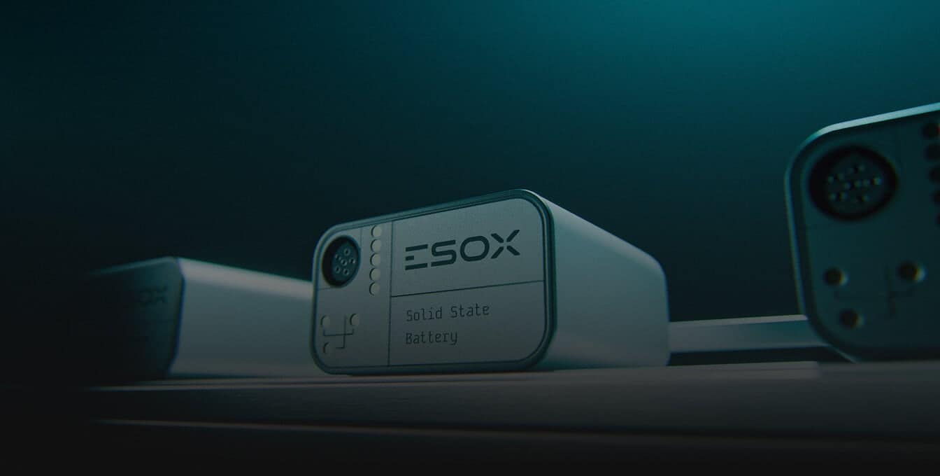 Close-up of a rectangular ESOX solid state battery with a plug connector, illuminated in a dark, teal-lit environment, sitting on a flat surface with part of another device visible beside it.