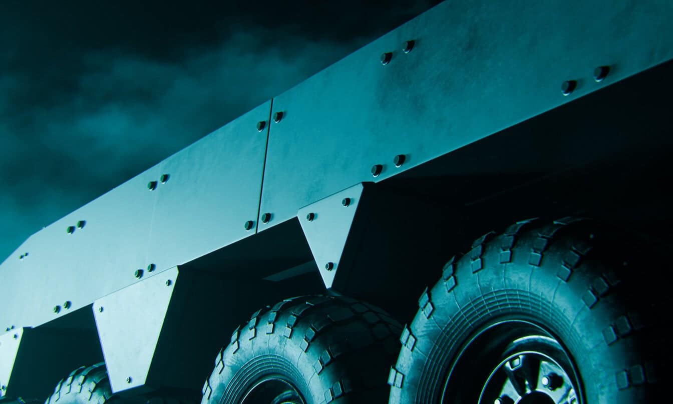Close-up, low-angle view of large armored vehicle wheels and metal-plated sides, illuminated by teal light against a dark, cloudy sky. The image emphasizes the rugged, industrial design and heavy-duty tires.