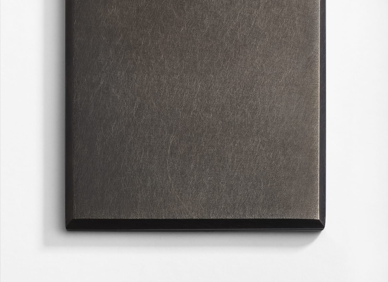 A close-up of a rectangular, brushed metal plaque with beveled edges, photographed on a white surface. The plaque has a smooth, reflective texture and a dark metallic finish.