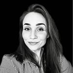 Black and white portrait of a woman with long hair, smiling softly. She wears a striped shirt and looks directly at the camera, against a solid black background.