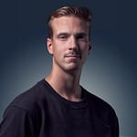 A young man with short, light brown hair and light facial hair wearing a black crew-neck shirt, posing against a gradient dark blue background.