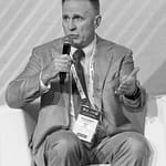 A man in a suit sits on a white chair, speaking into a microphone. He gestures with his left hand, wearing a conference badge around his neck. The image is in black and white.
