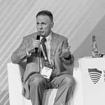 A man in a suit speaks into a microphone while seated on a stage at the World Police Summit. Two water bottles and event signage are on a table beside him.